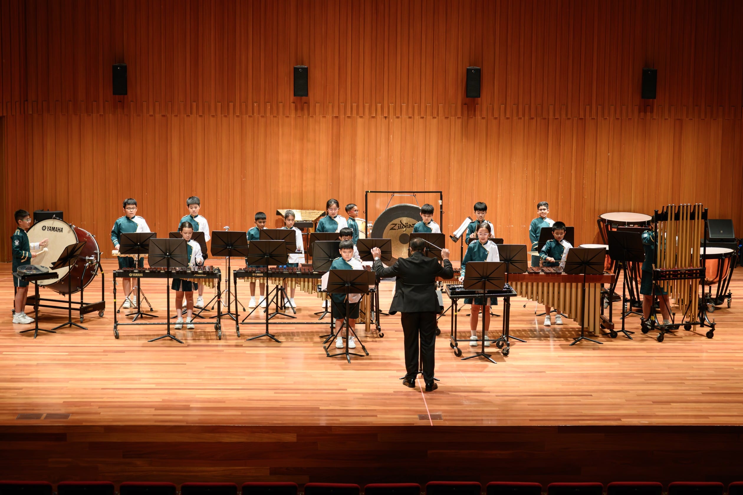 A wide-angle view of the school percussion ensemble performing on a concert hall stage, with students in band uniforms playing a full array of percussion instruments including bass drum, marimba, xylophone, gong, and timpani, led by a conductor facing the ensemble.